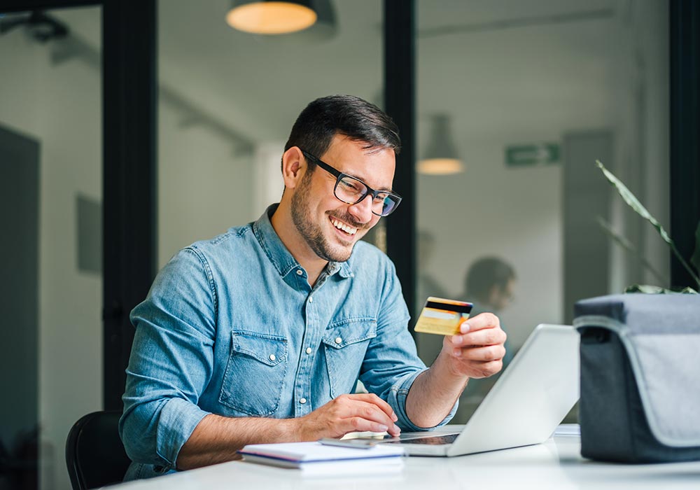 Photo of a smiling man, holding his credit card while shopping online with a laptop computer.