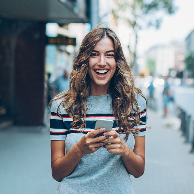Photo of a smiling woman standing on an urban street holding a smartphone. She is looking at the camera.